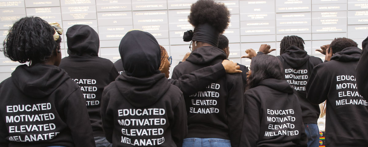 A group of Black students with their backs turned showing off sweaters that read “Educated, motivated, elevated, melanated”. 