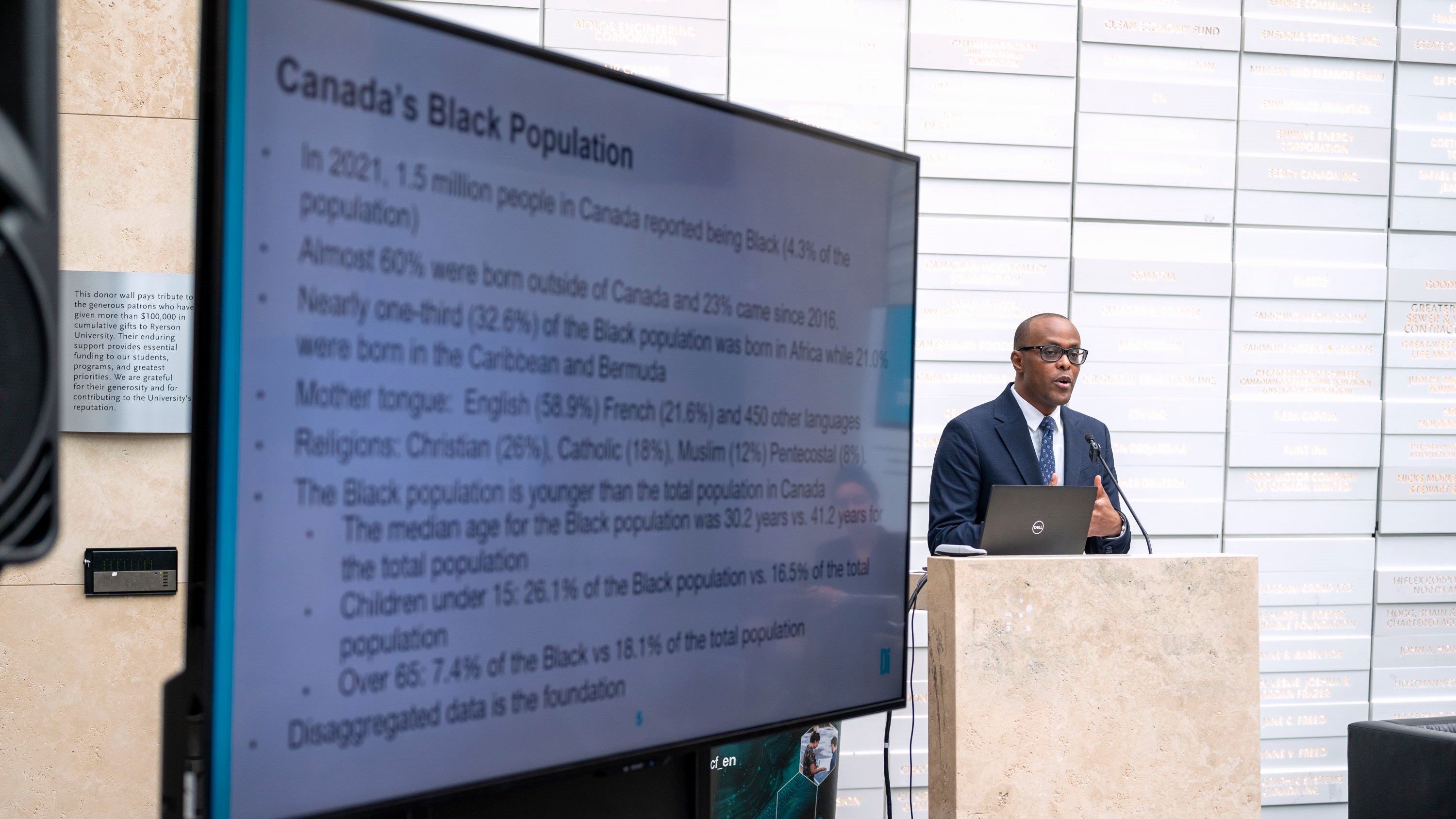 A Black man standing on a podium speaking, with a slide on the screen beside him that says Canada's Black Population.