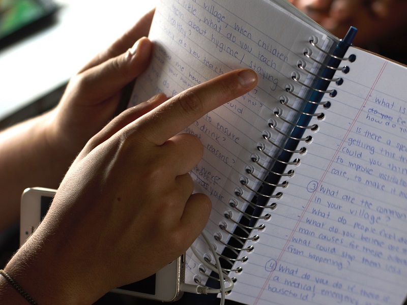 Hands hold and point to an open notebook with handwritten print. 