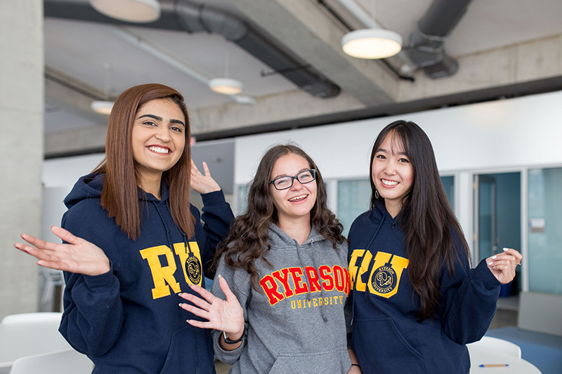Three students in Toronto Metropolitan University sweaters wave at the camera in the SLC.