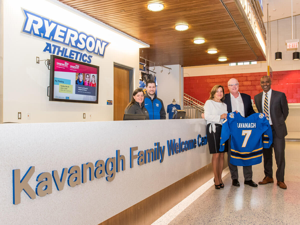 Three people holding a Rams jersey in front of the Ryerson Athletics desk at the MAC