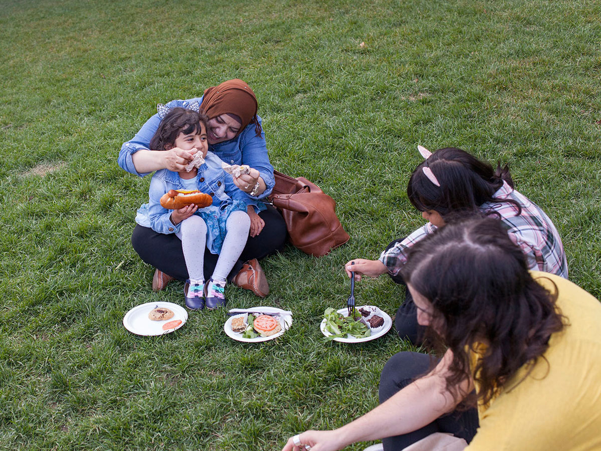 A mother feeding her child in her lap, having a picnic with her other children