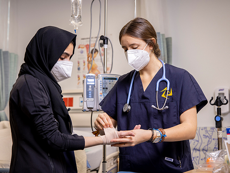 A nurse wearing a mask addressing a gauze bandage around a patient who is also wearing a mask in a medical facility.