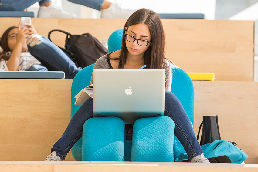 A student sits on a chair in the SLC, studying with a laptop in her lap. Another student on her phone can be seen in the background.