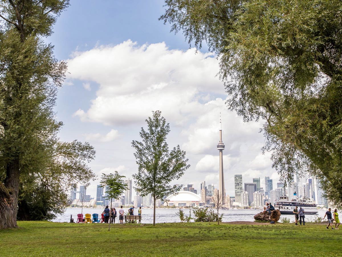 The Toronto skyline is seen across a body of water, in the foreground is grass, trees and people along the shoreline