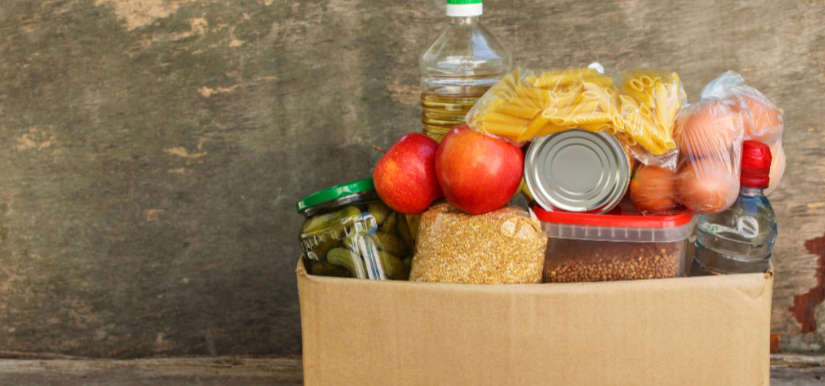 A brown box filled with food such as apples, a jar of pickles, eggs, dry pasta, and rice.