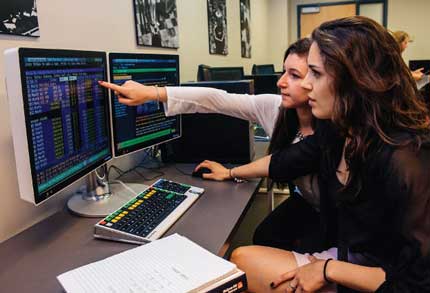 Two woman looking a computer screen, one of them is pointed to the data shown on the screen.