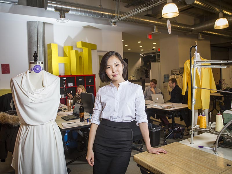 A fashion student stands and smiles while working in a fashion studio next to a sewing machine on a table and a mannequin with while fabric draped around it.