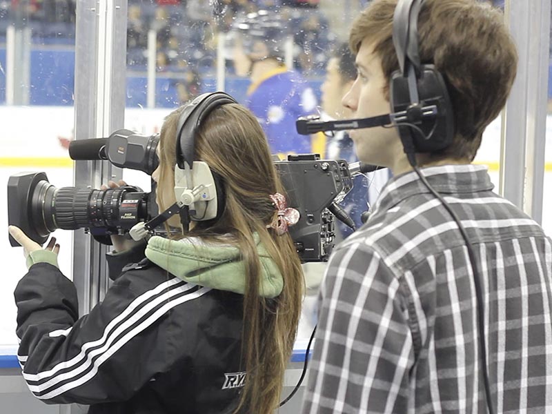 Two students, one holding a large television camera, and both wearing a headphone and microphone headset film a hockey game at the Mattamy Athletic Centre.