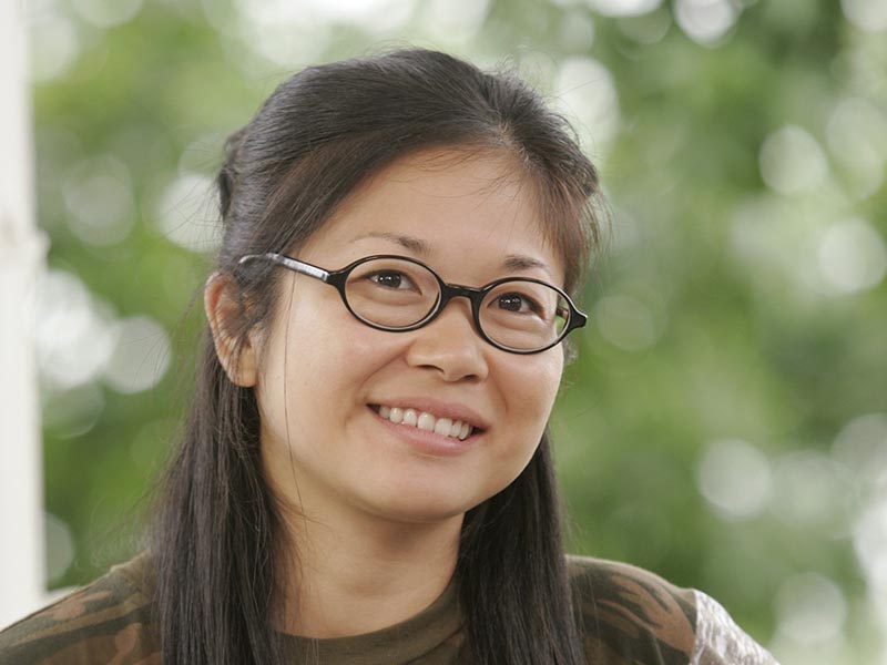 Lane Kim, portrayed by Keiko Agena, smiles in front of a light green background of leaves.