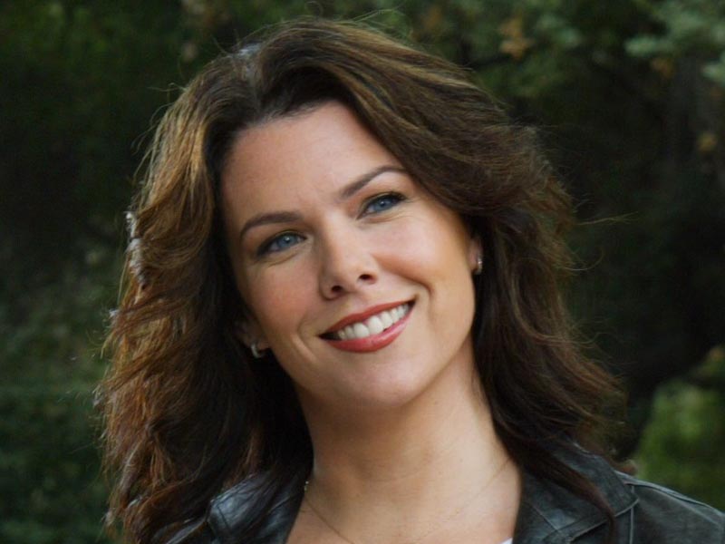Lorelai Gilmore, portrayed by Lauren Graham, smiles while looking away from the camera against a background of dark green leaves.