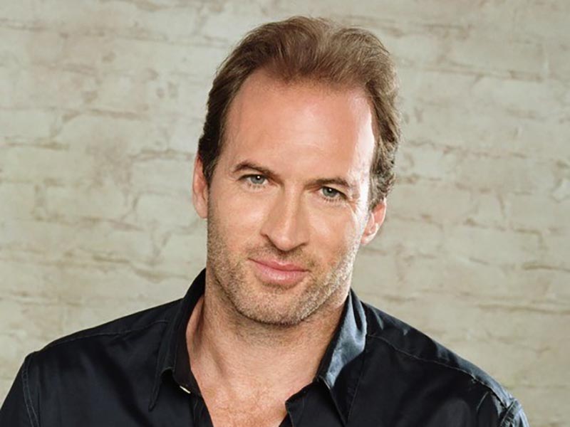 Luke Danes, portrayed by Scott Patterson, smiles against a white brick background.