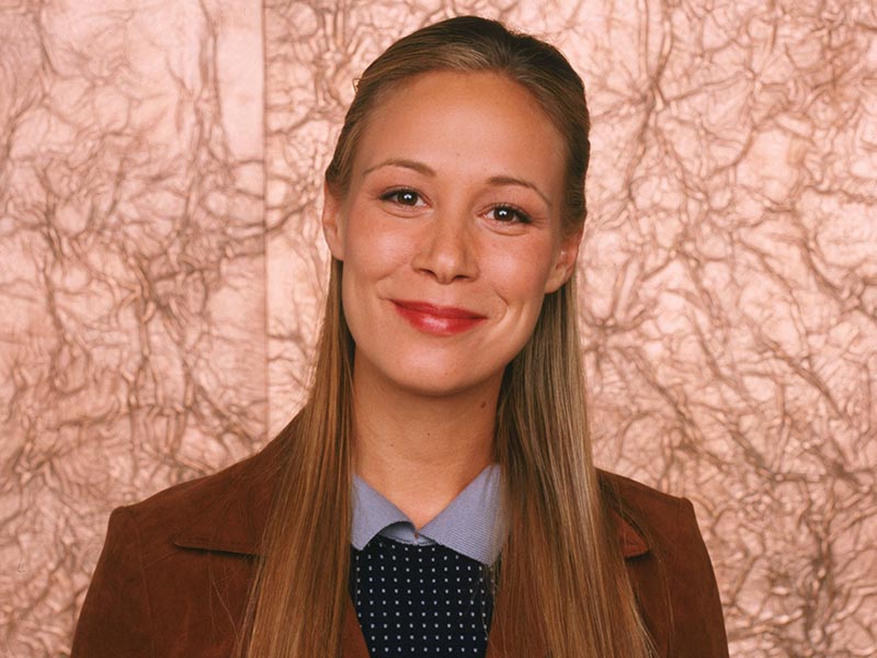Paris Gellar, portrayed by Liza Weil, smiles at the camera in a rust-coloured blazer against a pink marble background
