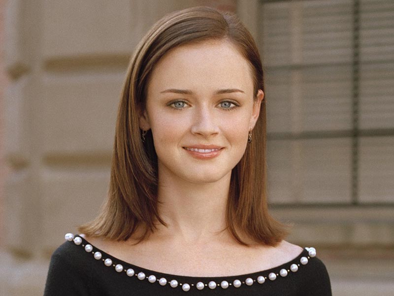 Rory Gilmore, portrayed by Alexis Bledel, smiles against a muted brown background of bricks and windows