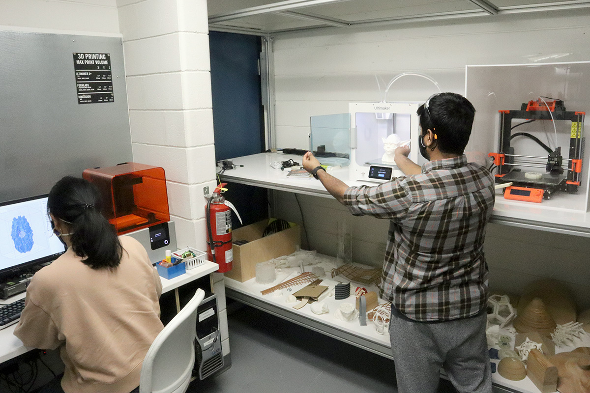 A student retrieves a small object from a 3D printing machine while another student works on a nearby desktop computer. 