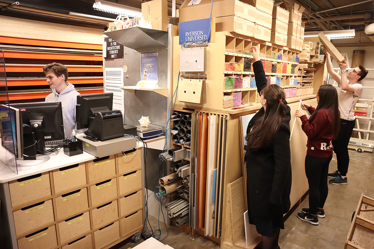 In the Material Store, three students browse dowels and other project materials located in a wooden shelving unit. A student staff member stands behind a checkout counter.