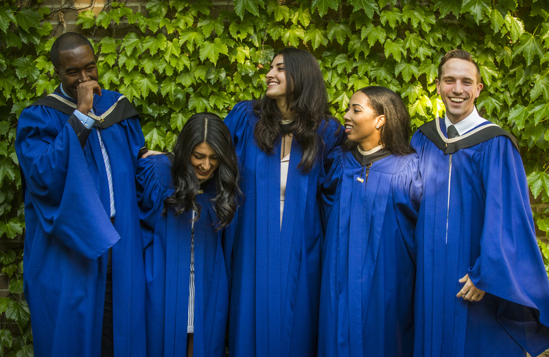 Five students stand together in front of a wall wrapped with vines, smiling in their convocation gowns.