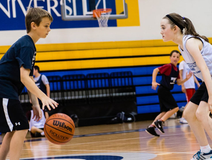 A boy dribbles a basketball in front of a girl who stands ready in defensive position