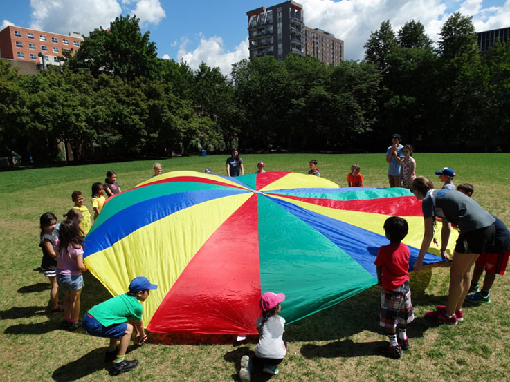 A group of children and camp counsellors pull a multi-coloured parachute to the ground, filling it with air