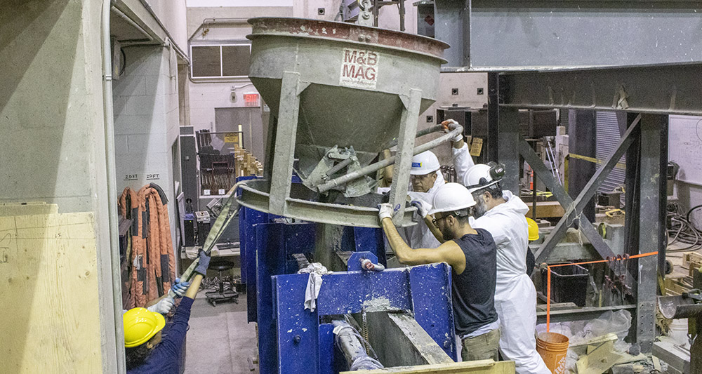 A group of researchers in a lab maneuver a large device that pours wet concrete into a cast