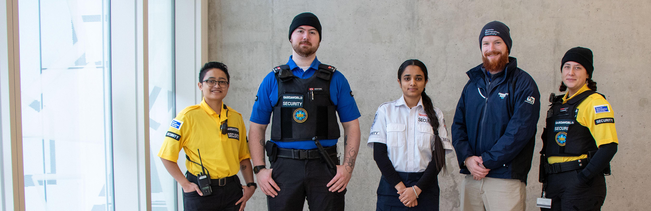 Five members of the Community Safety and Security team wearing the new uniforms and attire in front of a cement wall.