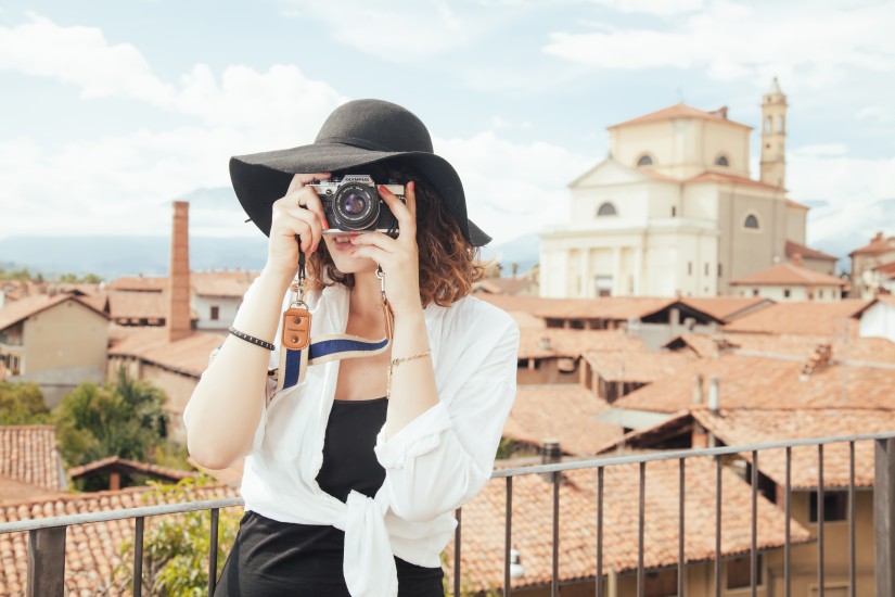 Woman photographer wearing a black hat is holding a camera with the backdrop of European town. 