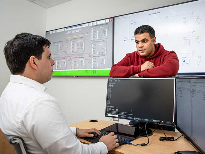 two researchers talking to each other in front of multiple monitors with electrical diagrams