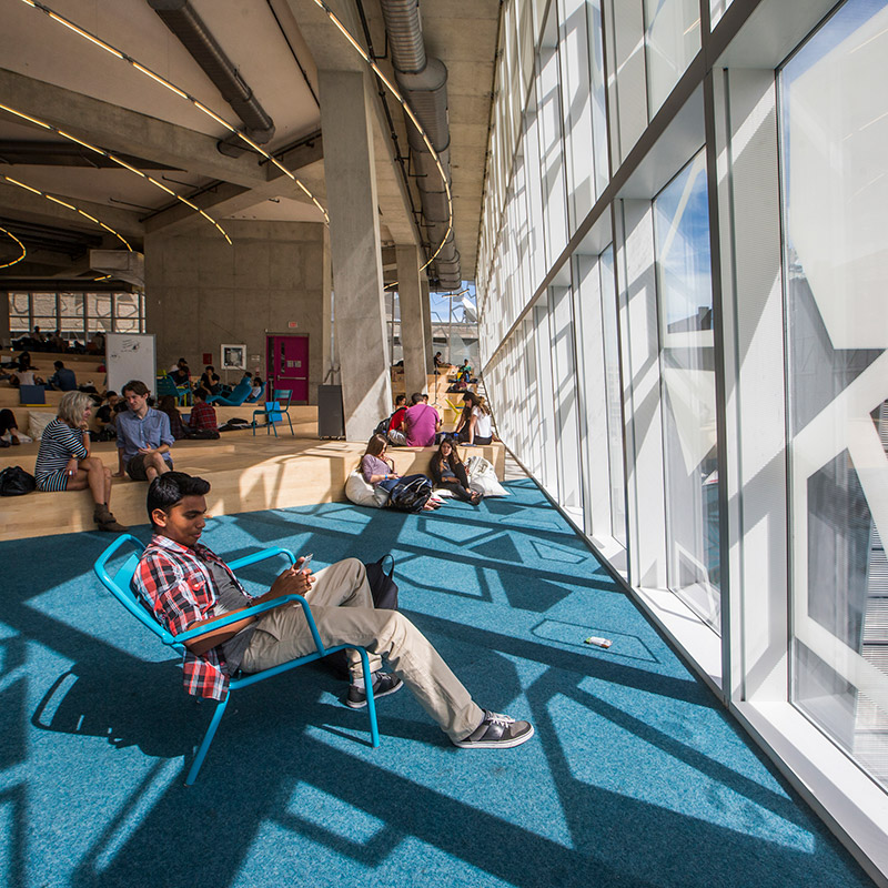 A student smiles as he checks his phone by the sunlit windows of the SLC.