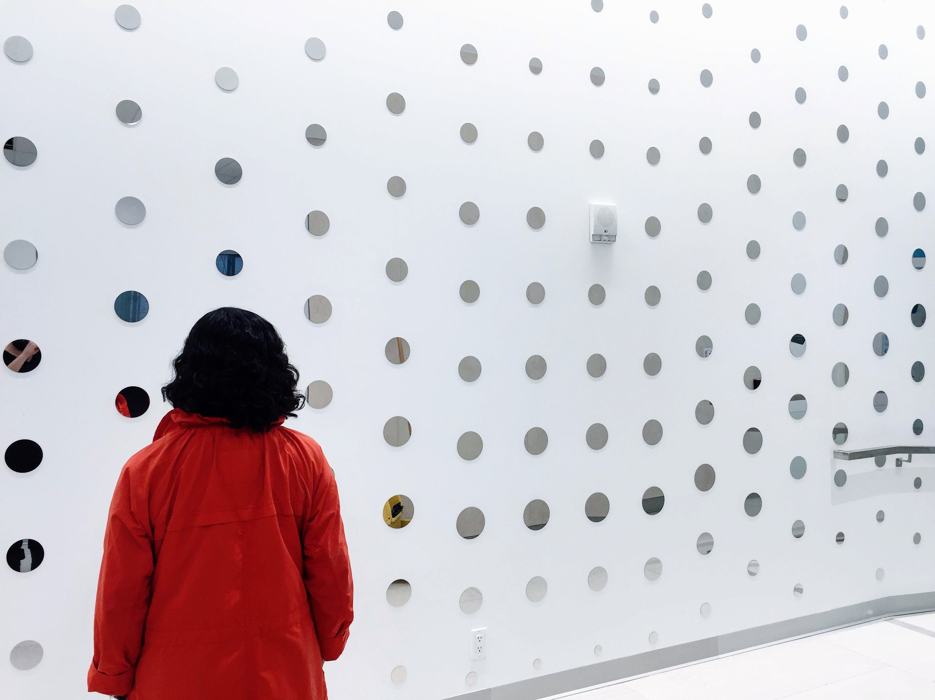 A figure stands in front of a white wall covered in  printed circles.