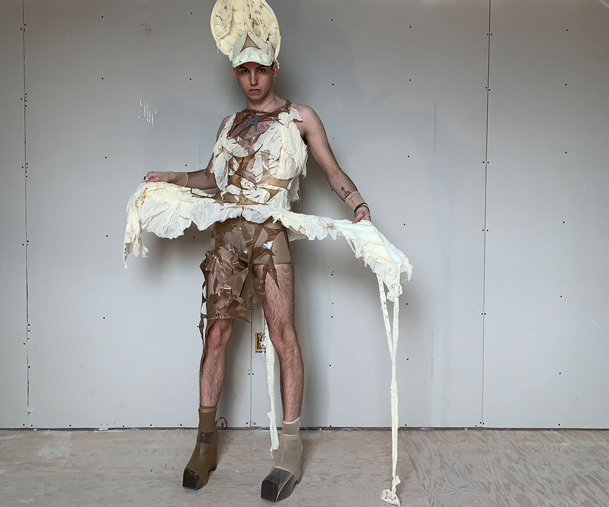 A model poses with a decorative white crochet garment. They wear a tall white headpiece and material dangles around their legs.