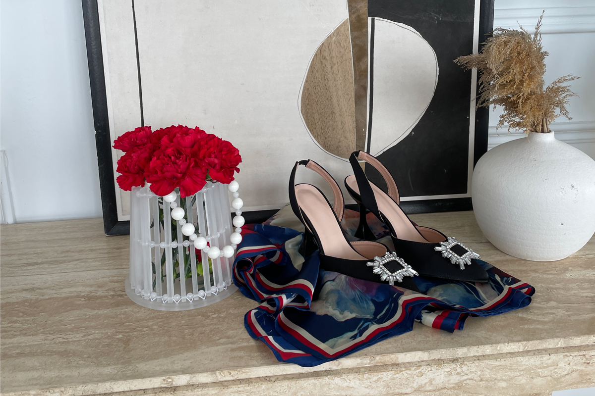 A series of objects on a surface. The objects include a cylindrical bag holding red flowers, a decorative scarf, a pair of black high heeled shoes and a white vase.