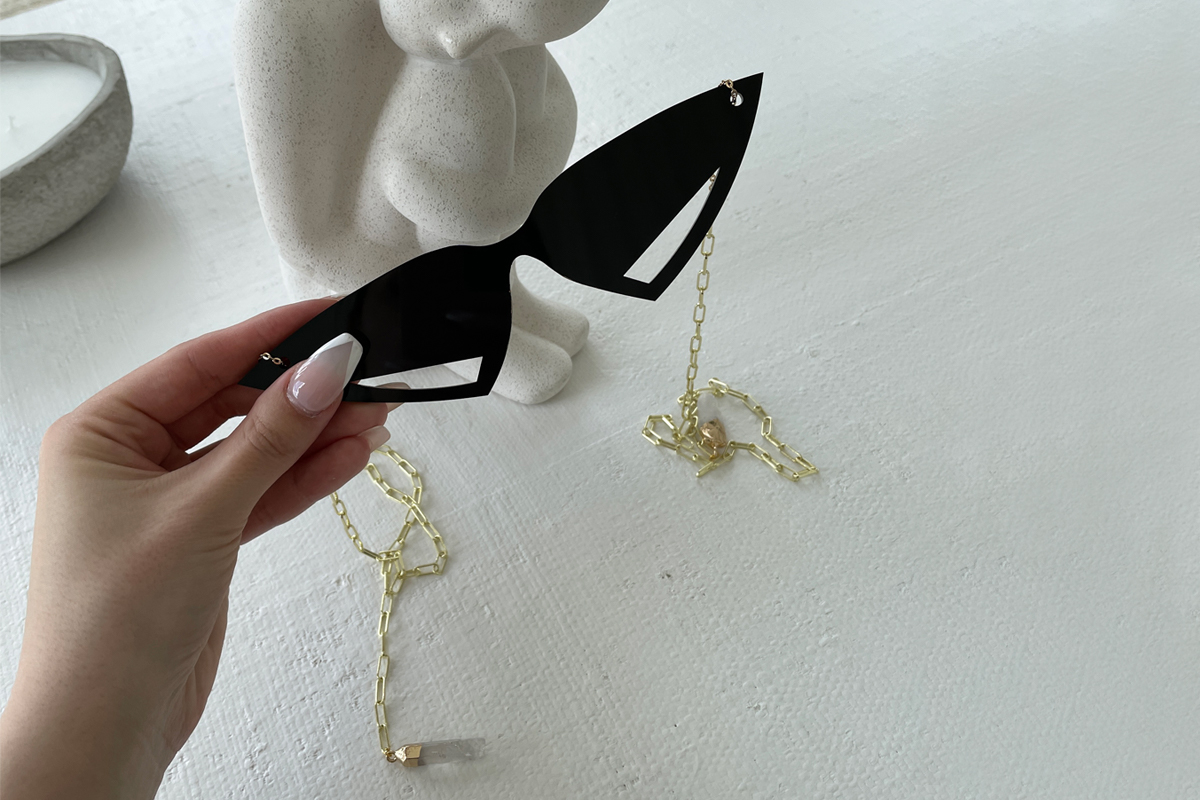Triangular sunglasses are held in hand, 2 gold chains drop to a table surface below.