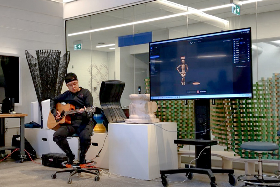 Someone wearing a motion-capture Suit sits down and plays guitar beside a screen showing a 3D model of a human figure in a similar position. There is lots of objects in the background.