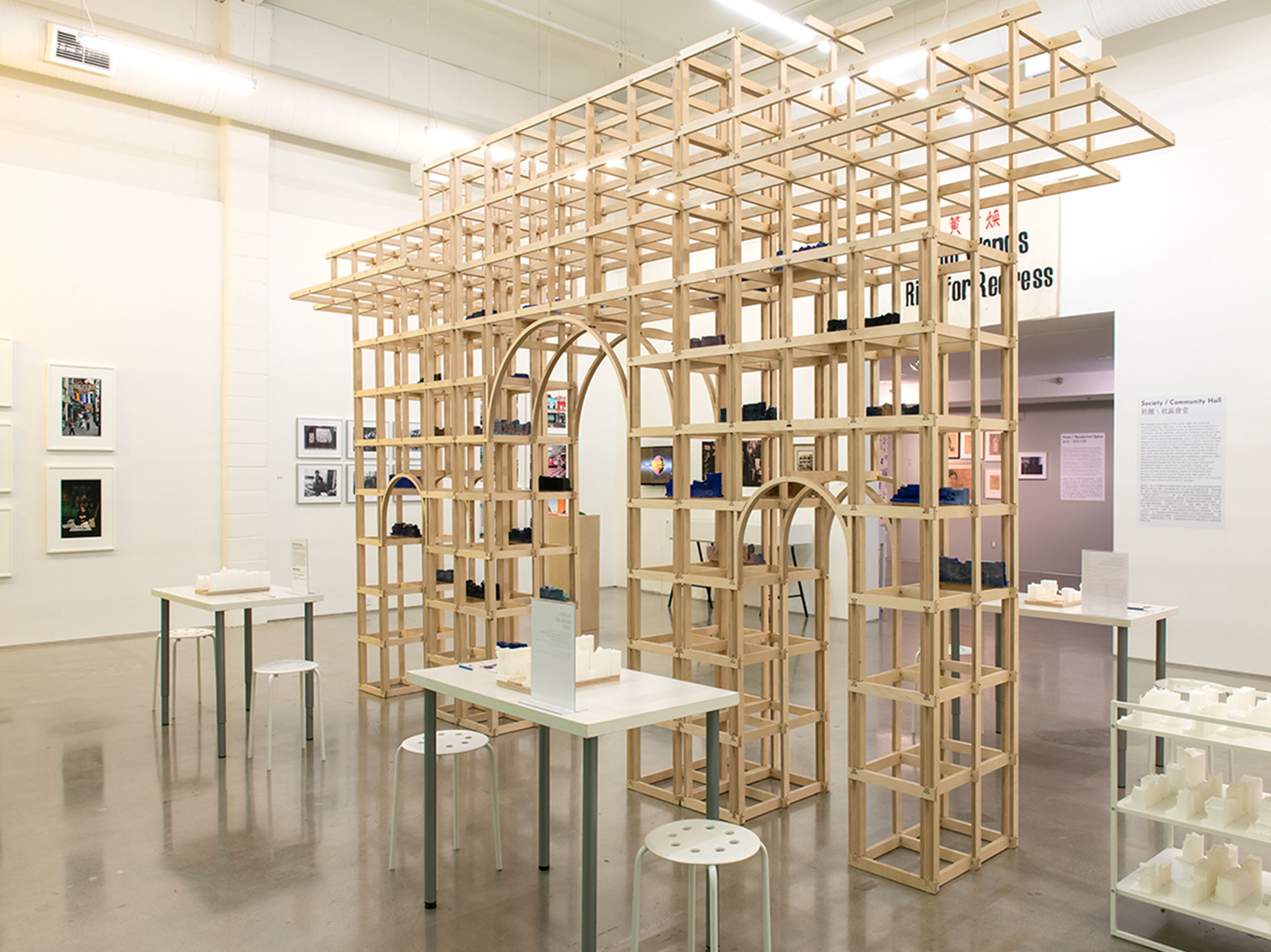 A gallery space with several objects on display. There is a wooden scaffold-looking structure, a tiered cart of small white objects, white tables with white stools and more white objects, and an adjacent room had framed art on the walls.