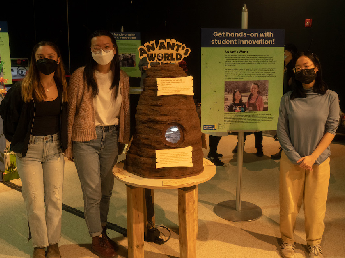 A student team stands proudly beside their anthill project in a dimly-lit room. An information poster for their project is displayed beside them on an easle. 