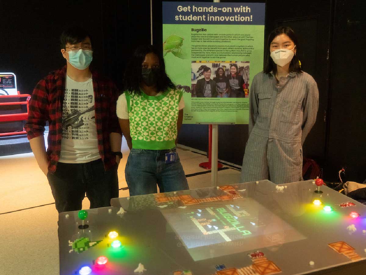 A student team stands proudly beside their project. There is a hand-made arcade table with multicolored lights and buttons. The students stand in front of an information poster of their project in a dimly-lit room.