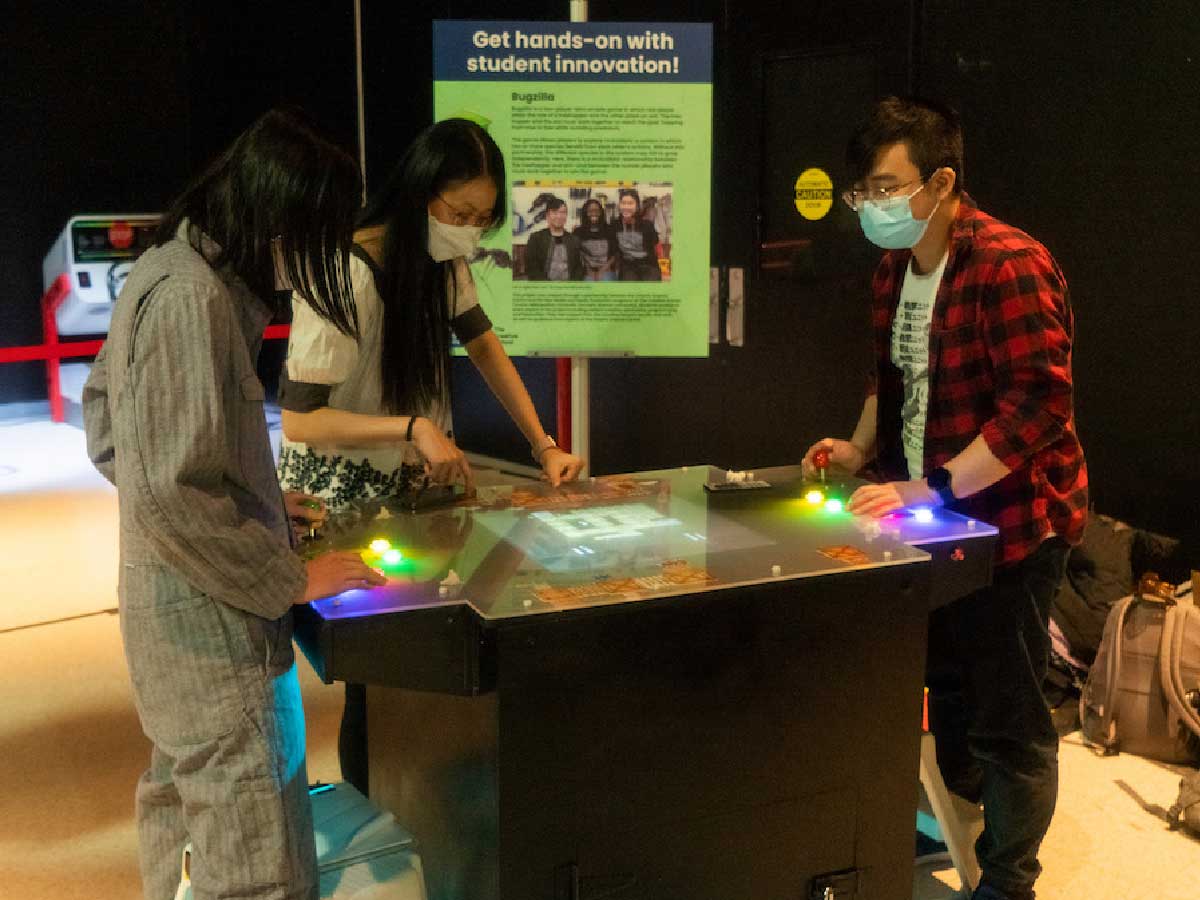 A group of students stand around a lit-up arcade table, playing the game.