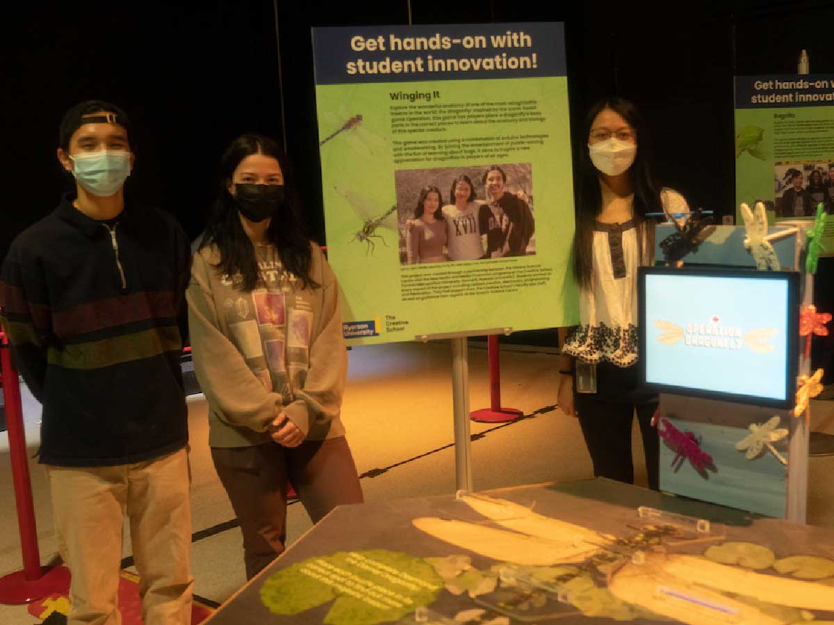 A student team stands proudly beside their project in a dimly lit room. Their project includes a wooden surface depicting a dragonfly, a screen surrounded by laser cut dragonflies. 