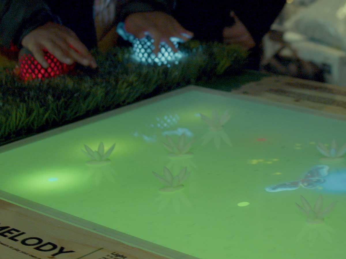 A student project features a glowing table surface depicting a pond. Someone's hand is hovering over glowing lanterns at the edge of the table.