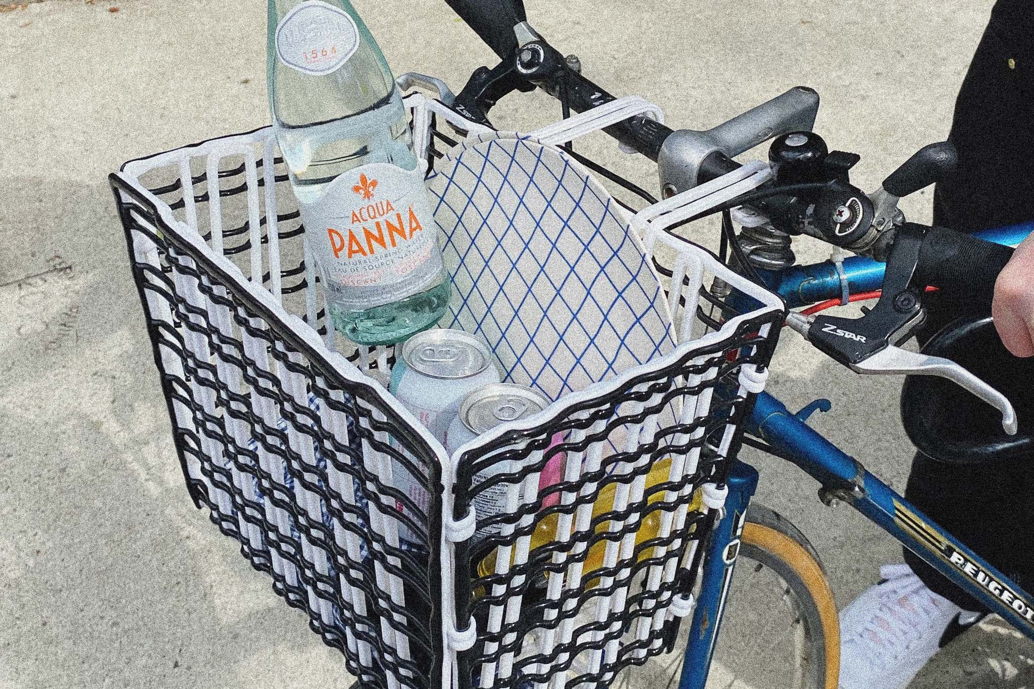 A close-up image of front bike basket on a bike. The basket is woven black and white plastic, resembling a textile. There are beverages and a paper plate inside the basket.
