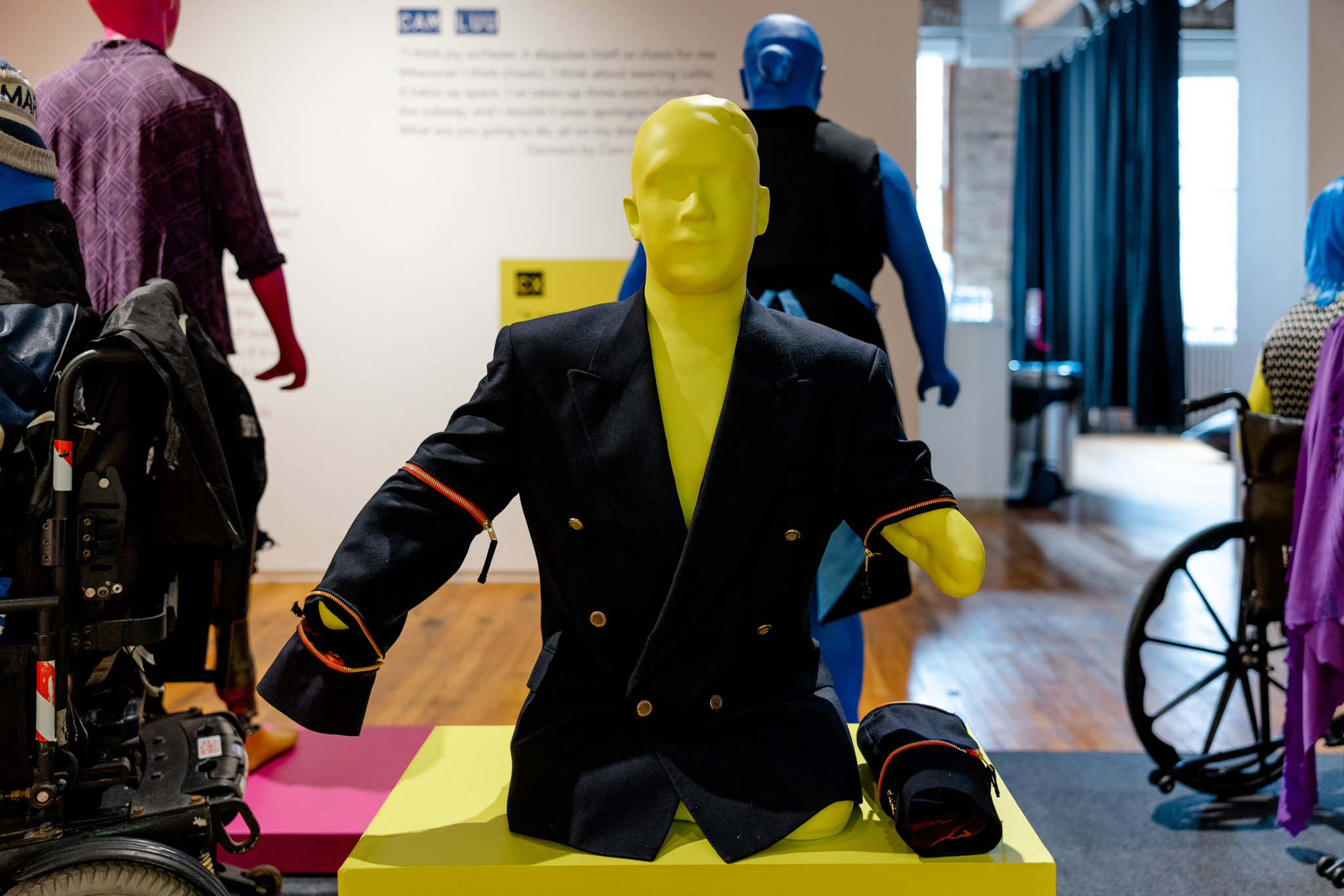 A 3D printed neon yellow mannequin wearing a navy blue suit jacket in a gallery setting. 
