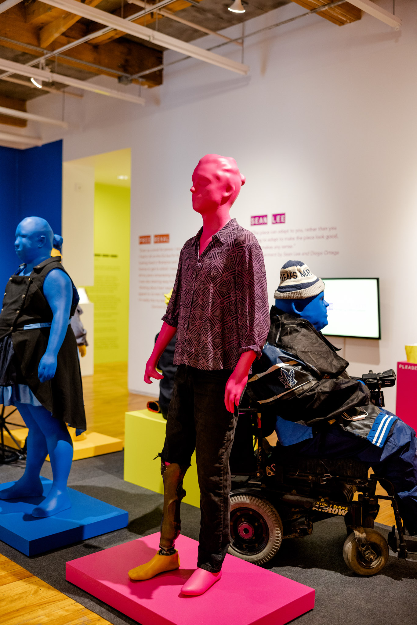 A bright pink mannequin stands in a gallery setting wearing a blousy top and jeans that show a prosthetic leg.