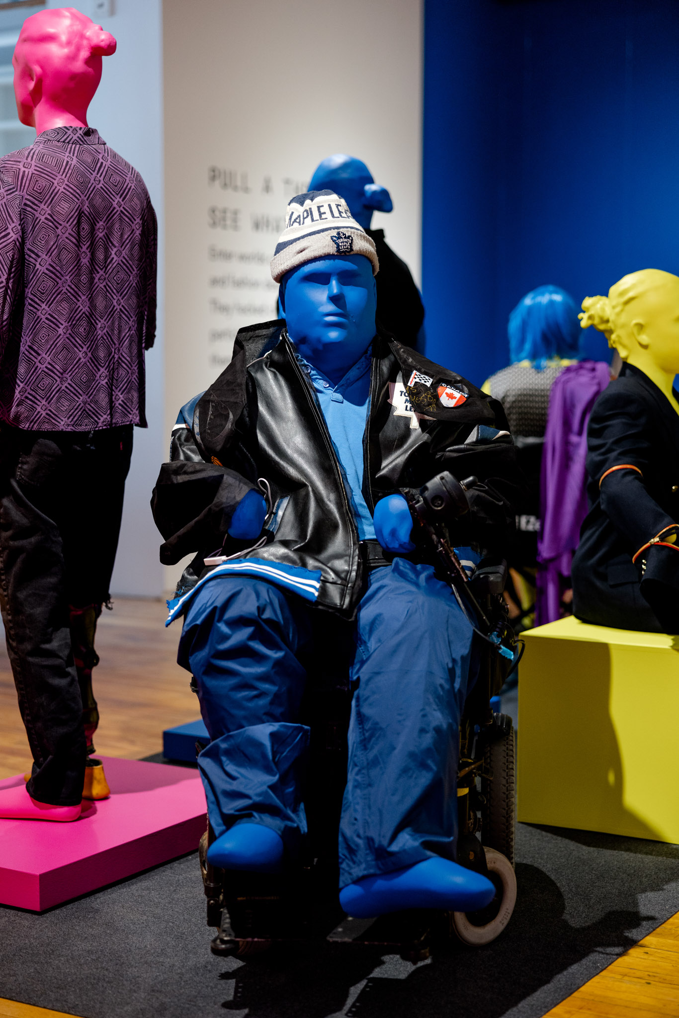 A Blue mannequin in a wheelchair is wearing a varsity jacket and toque in a gallery setting.