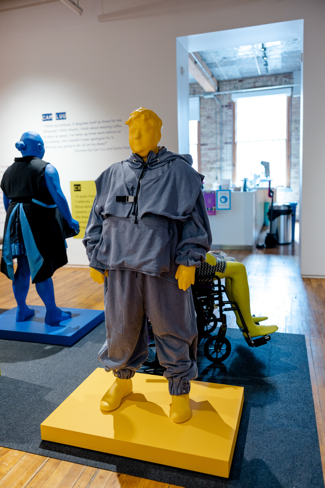 A bright yellow mannequin stands in a gallery setting wearing a blue jumpsuit. 