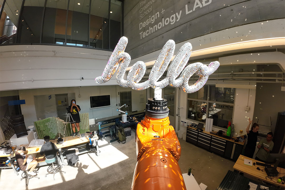 A large, orange Kuka robotic arm holds a 3D sculpture of the word "hello". The sculpture is fully coated in little reflective material pieces, mimicking a disco ball. There is a series of little light speckles across the wall behind the robot. 
