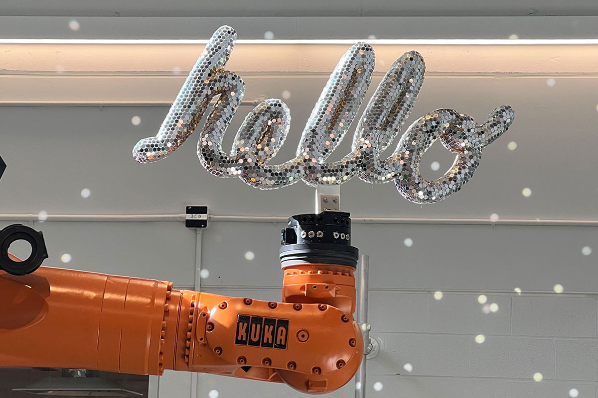 A large, orange Kuka robotic arm holds a 3D sculpture of the word "hello". The sculpture is fully coated in little reflective material pieces, mimicking a disco ball. There is a series of little light speckles across the wall behind the robot. 