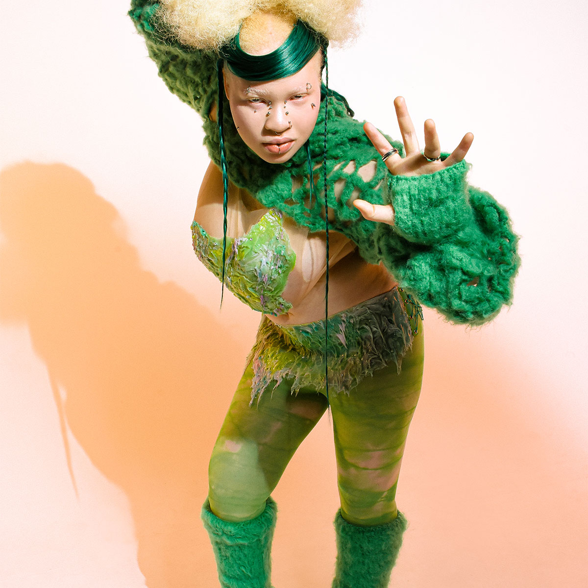 A model is wearing a green crochet shawl, shiny green headpiece, lacey green tights and knit green leg warmers. The model is leaning forward with one hand raised toward the camera.