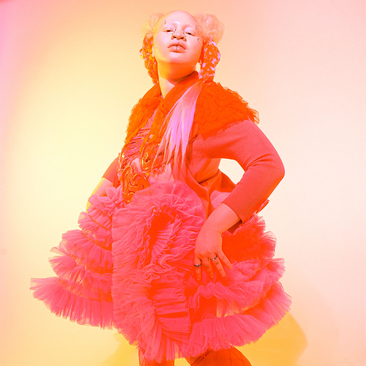 A model standing upright wearing a fluffy dress and shawl made of krinolin. The entire image is tinted red. 