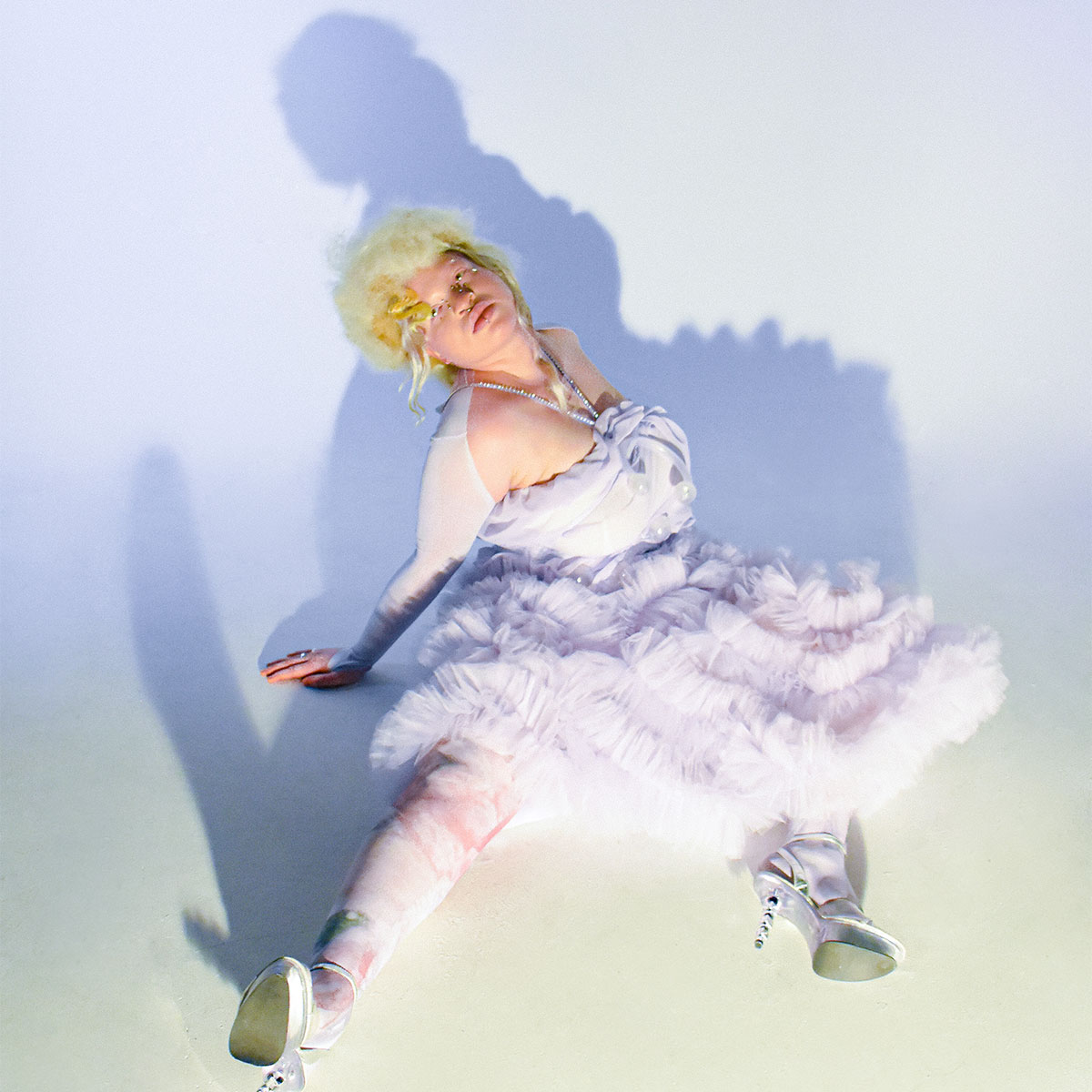 Model showcasing a poofy dress. The model is sitting on gthe ground leaning backwards with their legs apart. The dress is off-white, wearing multicoloured tights and white high-heeled shoes over a white photo backdrop.