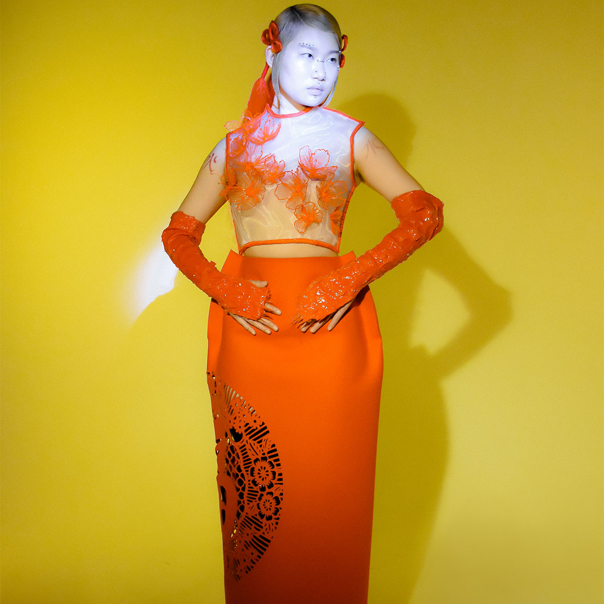 A model is standing up straight wearing a partially see-though decorative orange garment.  The garment is made of shiny material, with 3D flowers affixed, over a yellow photo backdrop. 